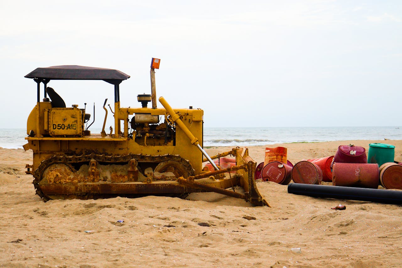 A rusty bulldozer and colorful barrels sit on a sandy beach by the sea, depicting coastal industrial work.